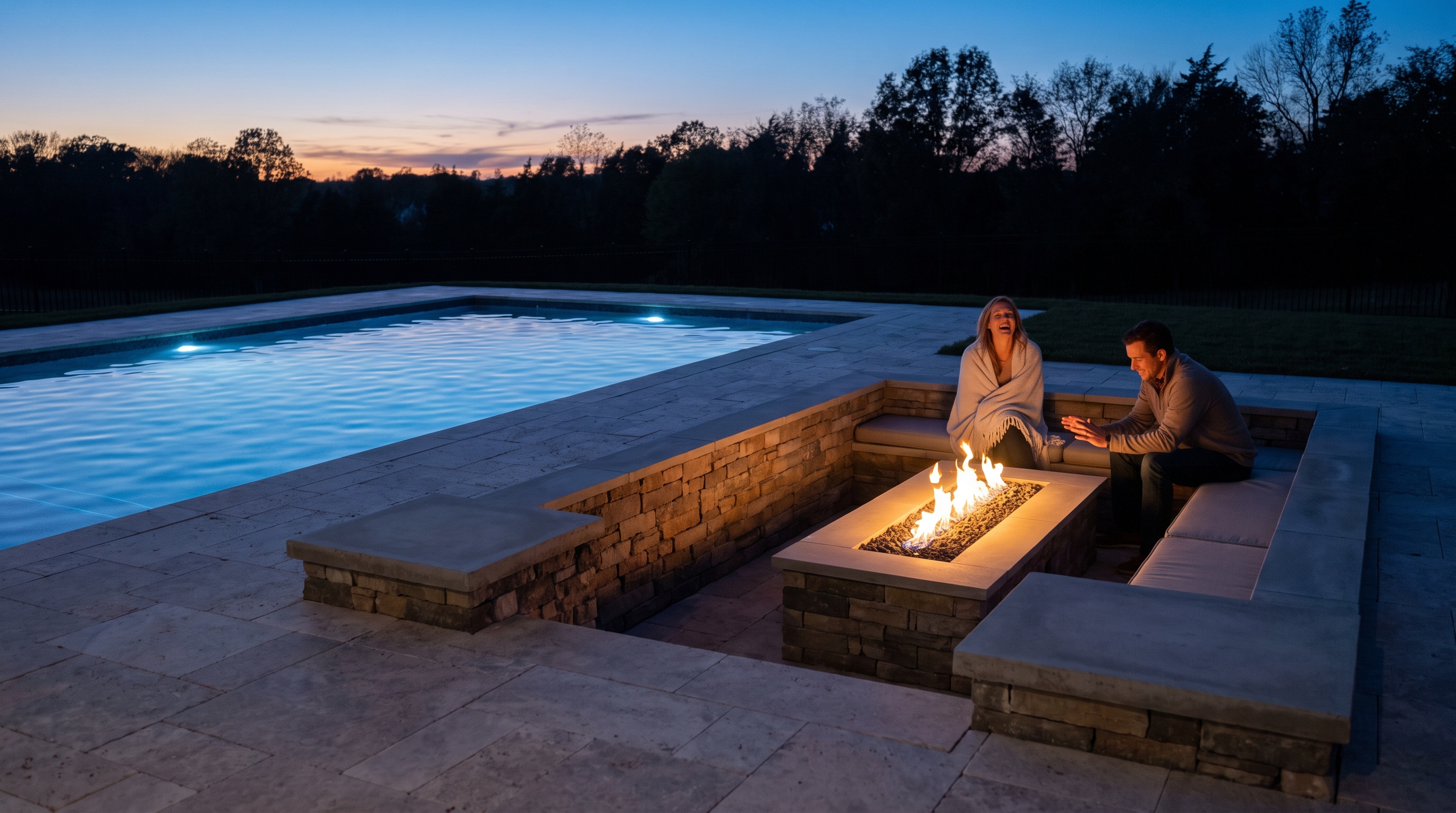Couple seated in a stone sunken fire pit lounge beside a lit pool at dusk